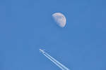 Ein Flugzeug zieht seine Kondensstreifen unterhalb des zunehmenden Mondes.
